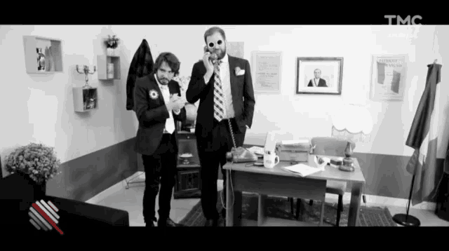 a black and white photo of two men talking on a phone with tmc written on the bottom right