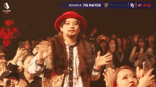 a man in a red hat stands in front of a crowd with the words round 8 7th match on the screen behind him