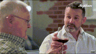 a man with a beard is holding a glass of red wine while talking to another man .