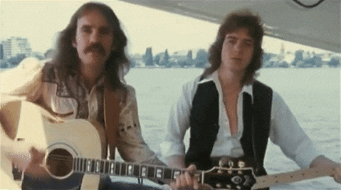 two men are playing guitars in front of a body of water with a city in the background