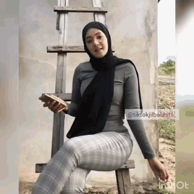 a woman in a hijab sits on a wooden ladder holding a cell phone