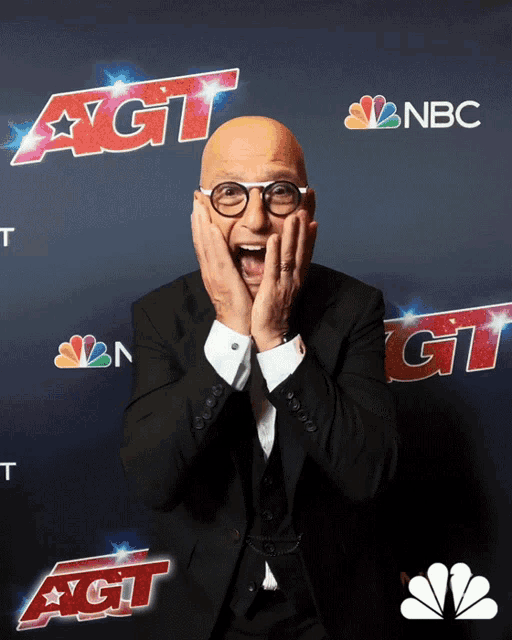 a bald man in a suit and glasses stands in front of a wall that says agt on it