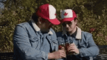 two men wearing canadian hats are sitting on a park bench toasting with beer .