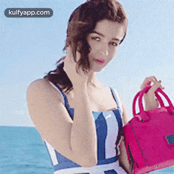 a woman in a blue and white striped dress is holding a pink purse in front of the ocean .