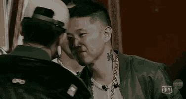 two men are standing next to each other and talking to each other . one of the men is wearing a hat .