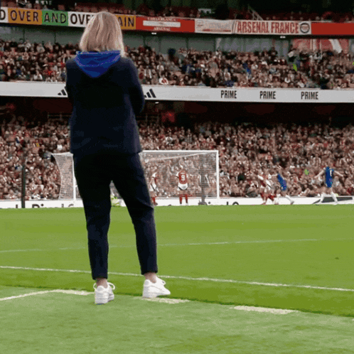 a woman stands on the edge of a soccer field watching a game