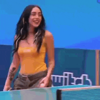 a woman in a yellow tank top and khaki pants is standing in front of a blue wall .