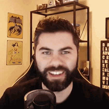 a man with a beard is smiling in front of a microphone in a room with wonder woman posters on the wall .