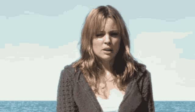 a woman in a grey sweater stands in front of a body of water
