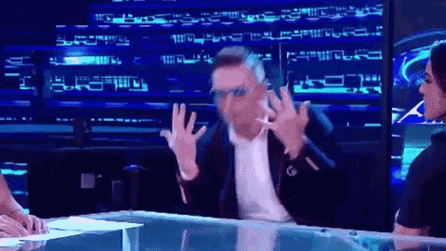 a man in a suit and sunglasses is sitting at a table with his hands up in the air .