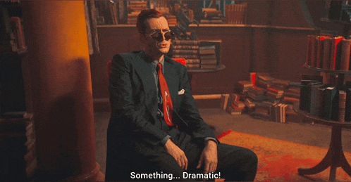 a man in a suit and tie is sitting in front of a bookshelf and says something dramatic