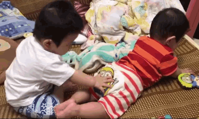 two young boys are playing with each other on a bed . one of the boys is wearing a minion shirt .