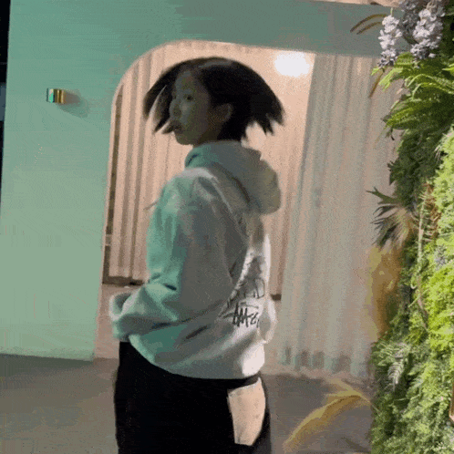 a woman wearing a stussy hoodie is standing in front of a green wall of plants