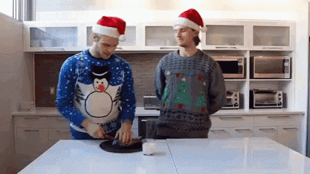 two men wearing santa hats and ugly christmas sweaters are standing in a kitchen preparing food .