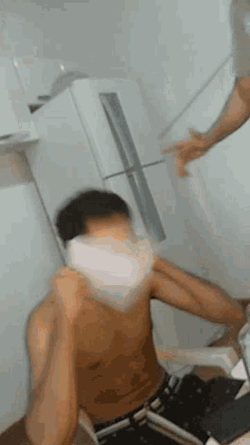 a shirtless man is sitting on the floor in front of a refrigerator while another man throws something at him .