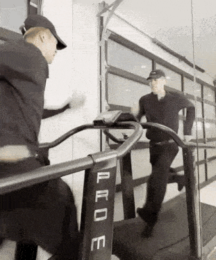 a man is running on a treadmill in front of a mirror while another man looks on .