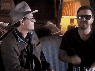 a man wearing a cowboy hat and sunglasses is talking to another man in a room .