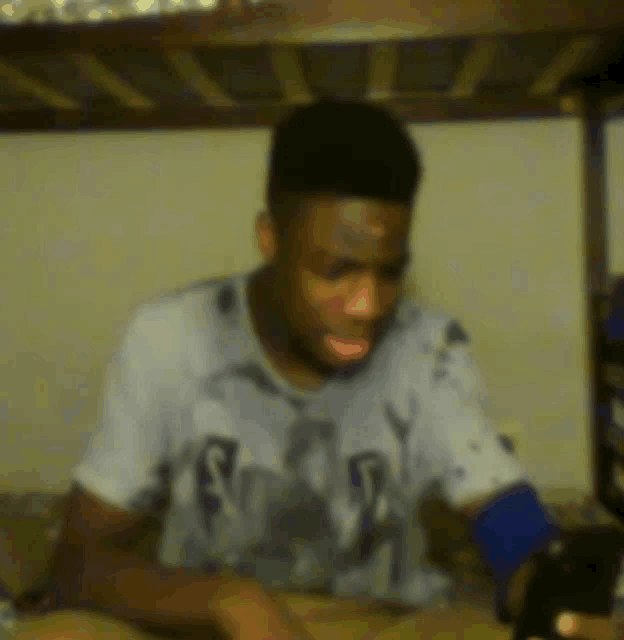 a man wearing a shirt with the letter r on it is sitting under a bunk bed