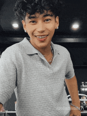 a young man with curly hair wearing a grey shirt and a necklace
