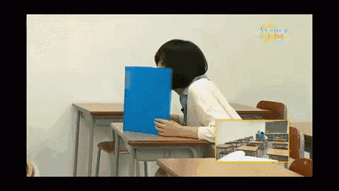 a woman is sitting at a desk in a classroom holding a blue folder in front of her face
