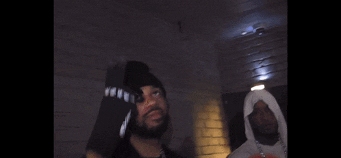 two men are standing next to each other in a dark room . one of the men is wearing a hooded jacket .