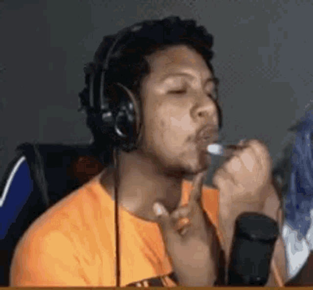 a man wearing headphones and an orange shirt is smoking a cigarette while sitting in front of a microphone .