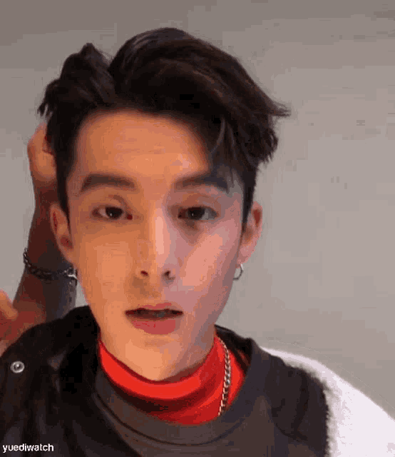 a young man wearing a red turtleneck and earrings is holding his hair .