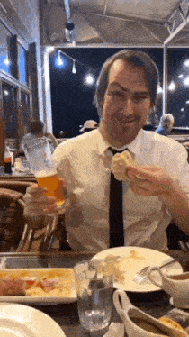 a man in a suit and tie is sitting at a table eating food and drinking a beer .