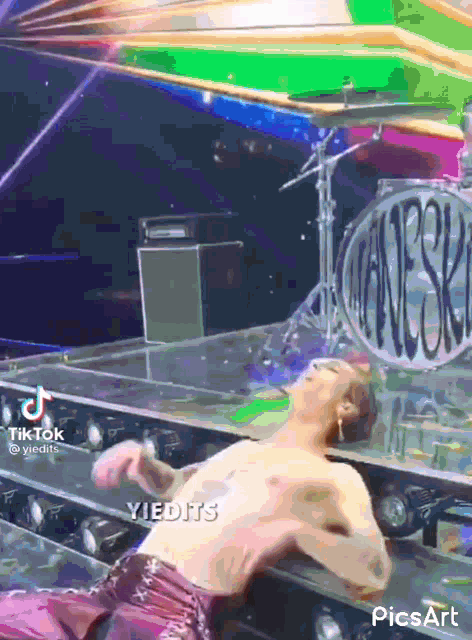 a man without a shirt is laying on a stage with a drum set in the background ..