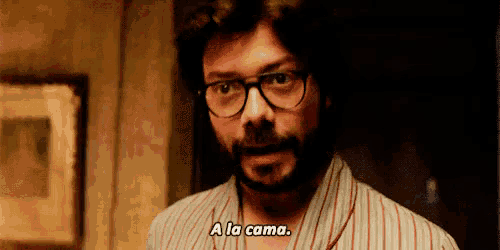 a man with glasses and a beard is standing in a dark room and says `` a la cama '' .