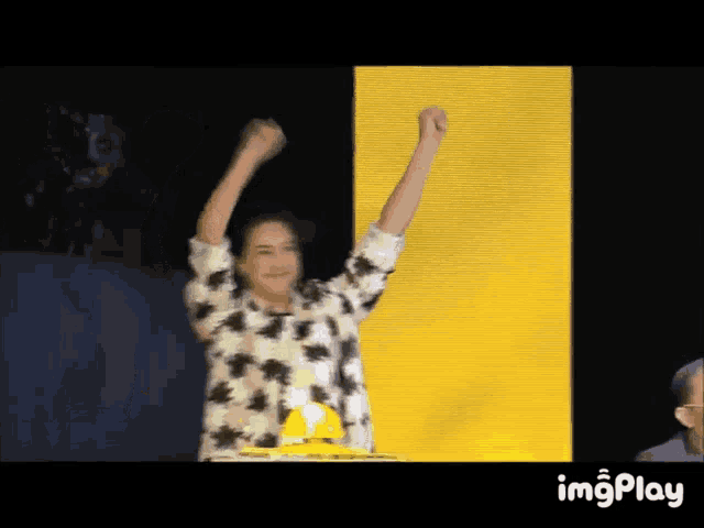 a woman in a floral shirt is raising her fist in the air in front of a yellow wall with imgplay at the bottom right