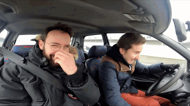 two men are sitting in a car with one covering his nose with his hand
