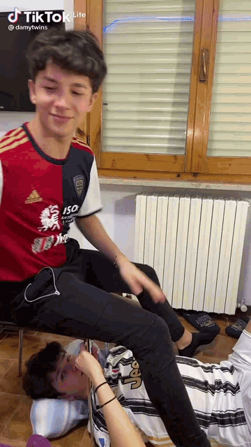 a boy in a red and white adidas shirt sits on another boy 's lap .