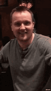 a man in a grey shirt is smiling and looking at the camera in a dark room .