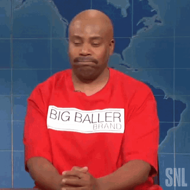 a bald man wearing a red shirt that says big baller brand is sitting with his hands folded .