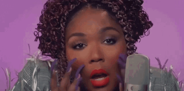 a woman with curly hair and purple nails is singing into a microphone on a purple background .