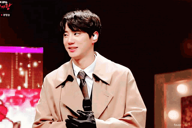 a man in a trench coat and tie is holding a microphone in front of a screen that says favor music