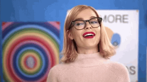a woman wearing glasses and a pink sweater is smiling in front of a colorful painting .