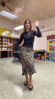a woman in a black shirt and a skirt is dancing in a classroom with a calendar on the wall