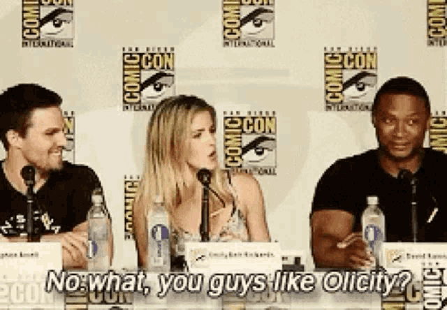 a group of people sitting at a table with bottles of water in front of a comic con wall
