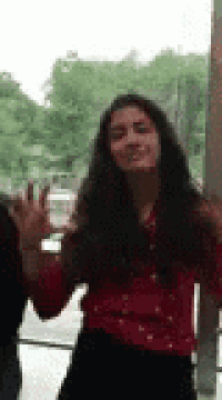a woman in a red shirt is standing in front of a window and waving .