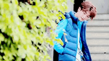 a young man wearing a blue vest is standing in front of a bush .