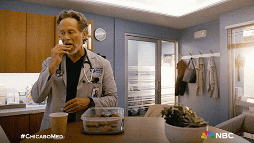 a man in a lab coat is sitting at a table with a nbc logo on the bottom right