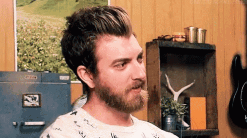 a man with a beard is sitting in a room in front of a shelf with a picture on it .