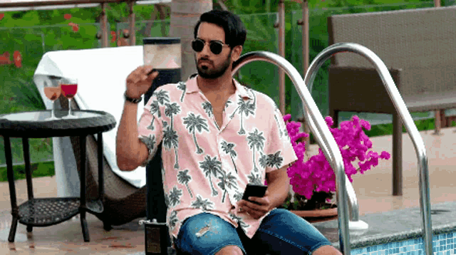 a man in a pink shirt with palm trees on it sits by a pool