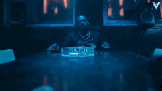 a man sits at a table in a dark room with an ashtray in front of him and the letter v on the wall
