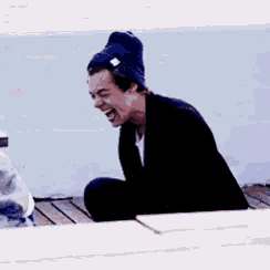 a man wearing a beanie is laughing while sitting on a wooden deck