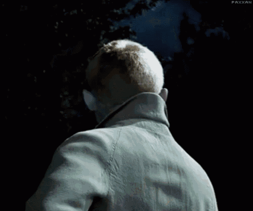 a man in a trench coat and tie is standing in the dark looking at the camera .