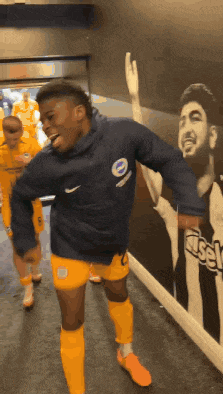 a soccer player wearing a blue hoodie and yellow shorts is dancing in a hallway