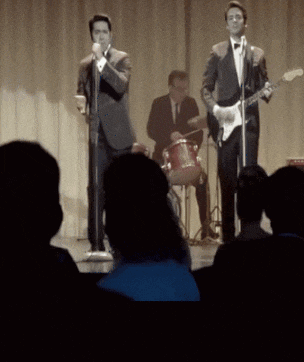 a man singing into a microphone with another man playing drums